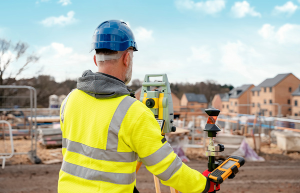 construction worker using surveying equipment outdoors wearing safety gear and hard hat focused on building site with homes and land development in background four key measurements