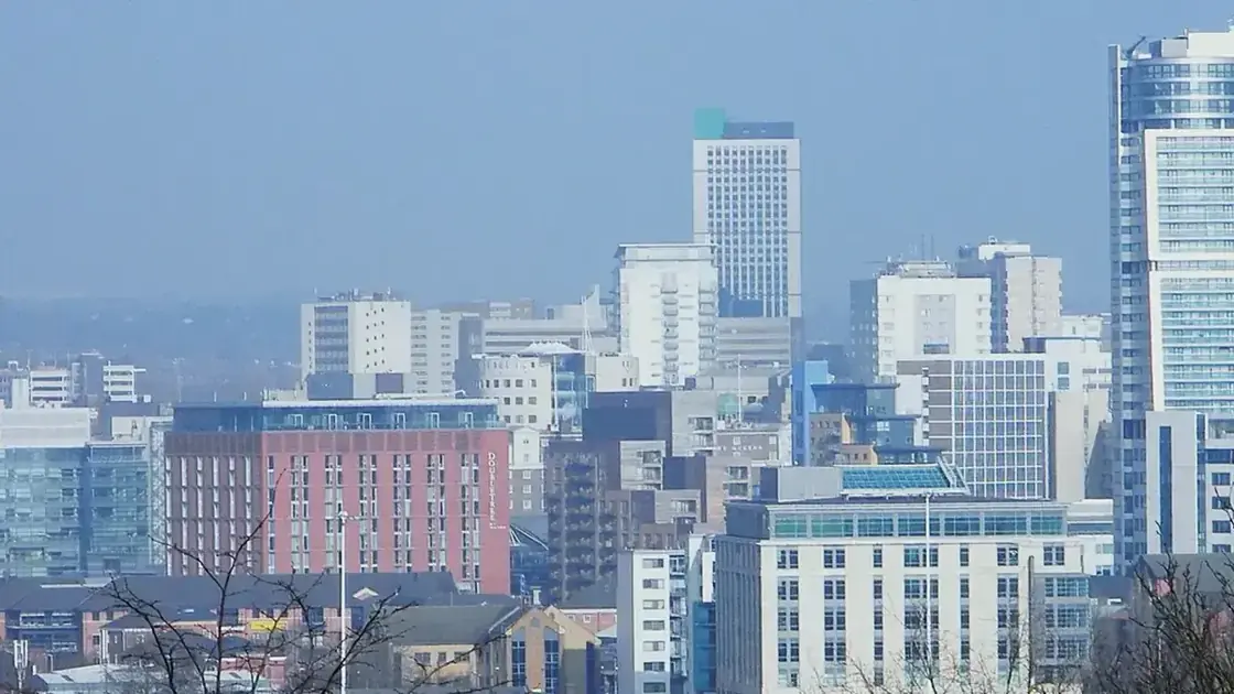 Wide daytime panorama of Leeds city-centre skyline from Beeston Hill, with rooftops in the foreground.